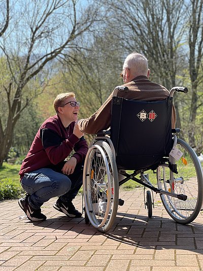 Pflegerin kniet vor älterem Mann im Rollstuhl